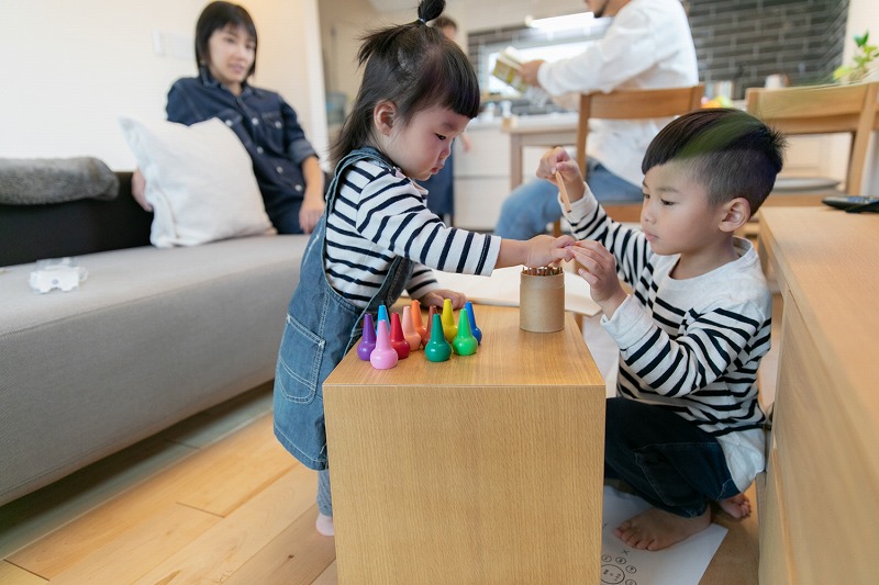 建築工房いろはの注文住宅のリビングで、小さなきょうだいが木製テーブルで色鉛筆やおもちゃを使って遊び、後ろで家族がくつろいで過ごす様子を写した写真で、家づくりや工務店選びの参考になる生活シーン。