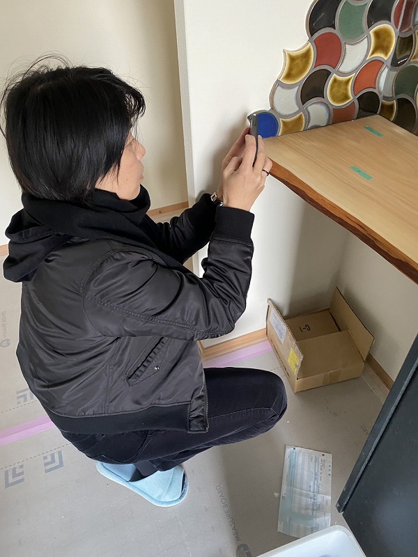 注文住宅の現場で、建築工房いろはのスタッフが造作棚とカラフルなタイル仕上げをスマートフォンで記録している様子を撮影した写真で、家づくりにおける内装確認と施工品質のチェックを示している。