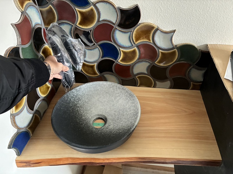 注文住宅の施工現場で、建築工房いろはが造作した木製カウンターに設置予定の陶器製手洗いボウルと黒色水栓を確認する様子が写され、家づくりにおける造作手洗いとタイルデザインのこだわりが伝わる。