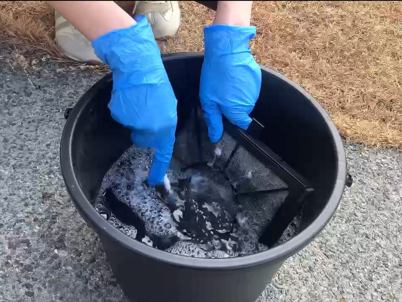 ALTテキスト（社名オプション：建築工房いろは）：
建築工房いろはが家づくりや注文住宅のメンテナンス工程として、青い手袋を着用した手が屋外でバケツの洗浄液に浸したフィルターをブラシでこすり洗いしている様子を写した写真で、住宅会社や工務店の点検作業を客観的に示す内容。