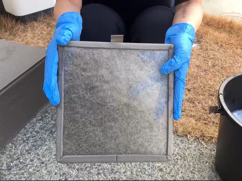 ALTテキスト（社名オプション：建築工房いろは）：
建築工房いろはが家づくりや注文住宅のメンテナンス工程として、青い手袋を着用した手が屋外でバケツの洗浄液のそばでホコリの付着したフィルターを持ち確認している様子を写した写真で、住宅会社や工務店の点検作業を客観的に示す内容。