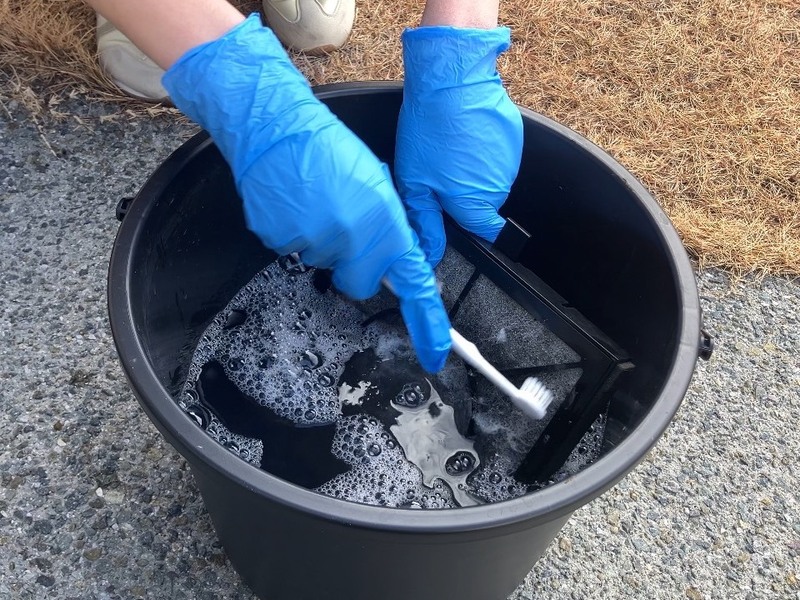 青い手袋を着用した人物が屋外で黒いフィルターをブラシで洗浄している様子を写した写真で、建築工房いろはによる住宅設備のメンテナンスや家づくりに関わる清掃作業を示す内容。