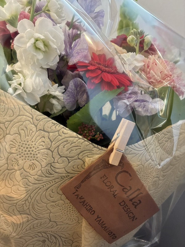 透明フィルムに包まれた花束に白いストックや赤いガーベラ、カーネーションなどがアレンジされ、木製クリップでフローラルデザインのタグが留められた様子を写した写真で、建築工房いろはの家づくりや注文住宅の引き渡しシーンにも使われる贈り物としてのイメージを伝える内容。