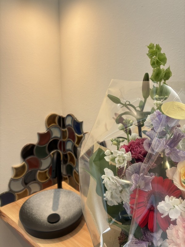 透明フィルムに包まれた花束に白いストックや赤いガーベラ、カーネーションなどがアレンジされ、木製クリップでフローラルデザインのタグが留められた様子を写した写真で、建築工房いろはの家づくりや注文住宅の引き渡しシーンにも使われる贈り物としてのイメージを伝える内容。