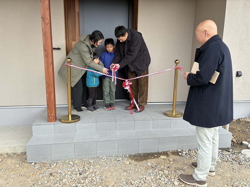 建築工房いろはが手がけた注文住宅の前で家族がテープカットを行う様子を写した写真で、工務店による家づくりのお引渡しの記念シーンを捉えている。