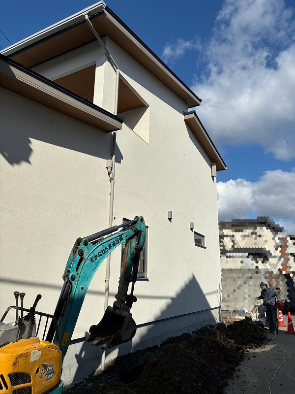 建築工房いろはが施工する注文住宅の敷地で、小型重機が建物横の地面を掘削し配管工事を進める様子を写した写真で、家づくりや工務店の現場管理の一環として作業員が丁寧に確認しながら進めている状況を客観的に示した内容。