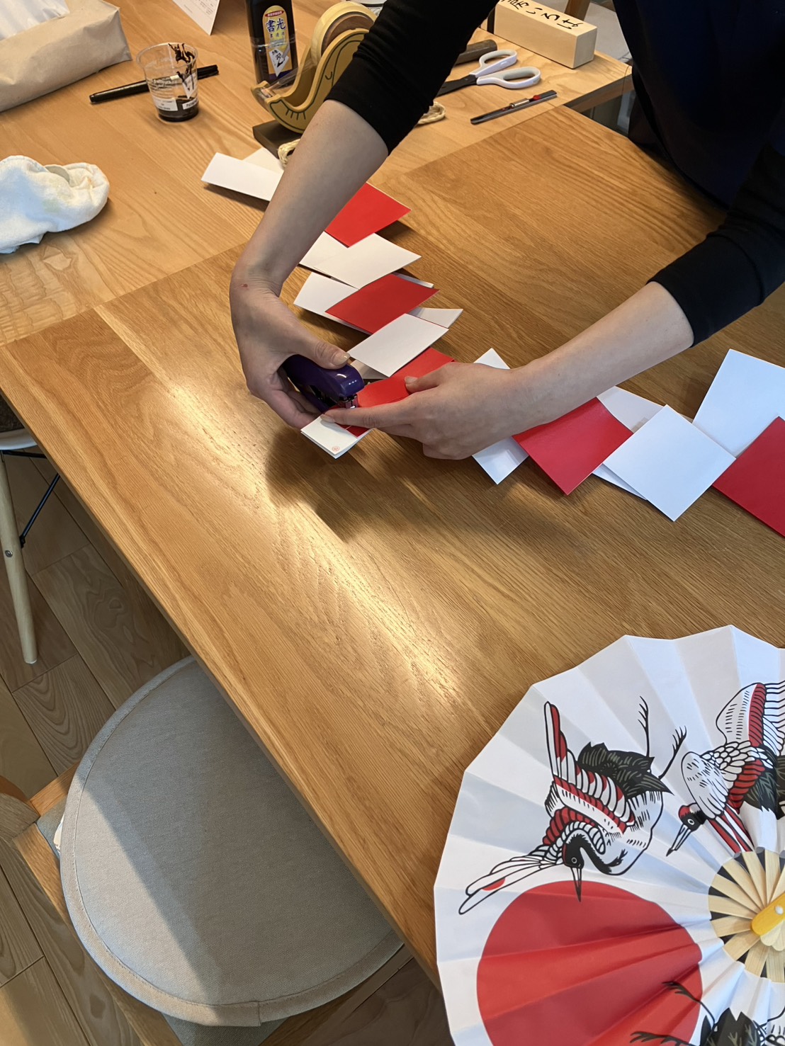 ALTテキスト(社名オプション:建築工房いろは):
木目調のテーブルの上で、建築工房いろはのスタッフが赤と白の紙を交互に繋ぎ合わせる作業を行っており、手前にはおめでたい鶴の絵が描かれた祝い扇子が置かれている、注文住宅の地鎮祭や上棟式に向けた式典準備の様子。