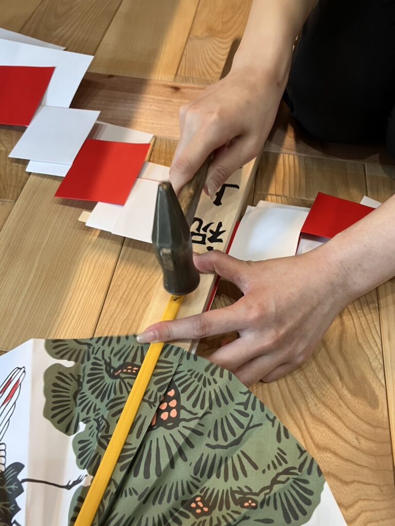 建築工房いろはのスタッフが、上棟式で使われる幣束に縁起物の扇を金槌で丁寧に固定している様子で、無垢材のフローリングの上で注文住宅の完成と工事の安全を願って一つひとつ手作業で準備を進める工務店のこだわりが伝わる写真。