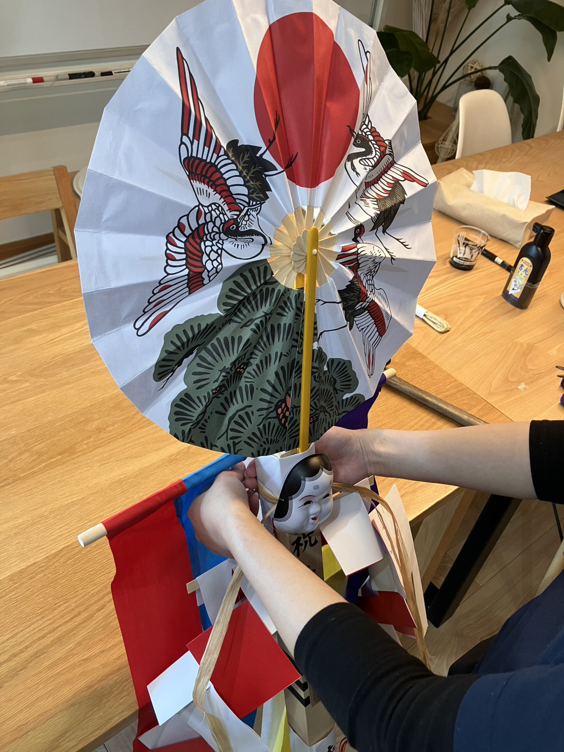 建築工房いろはのスタッフが、日の出や松、つがいの鶴が描かれた縁起の良い扇飾りとおかめの面を幣束(へいそく)の先端に丁寧に取り付けている、注文住宅の上棟式に向けた準備風景であり、家づくりの安全とご家族の繁栄を願う工務店の手仕事が伝わる写真。