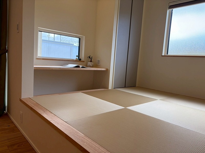 建築工房いろはによる注文住宅の施工事例で、リビングの一角に設けられた小上がりの畳コーナーには、読書や作業に便利な造作デスクと、柔らかな光を採り込むスクエア型の窓が配置された、機能的で落ち着きのある和モダンな空間の写真。