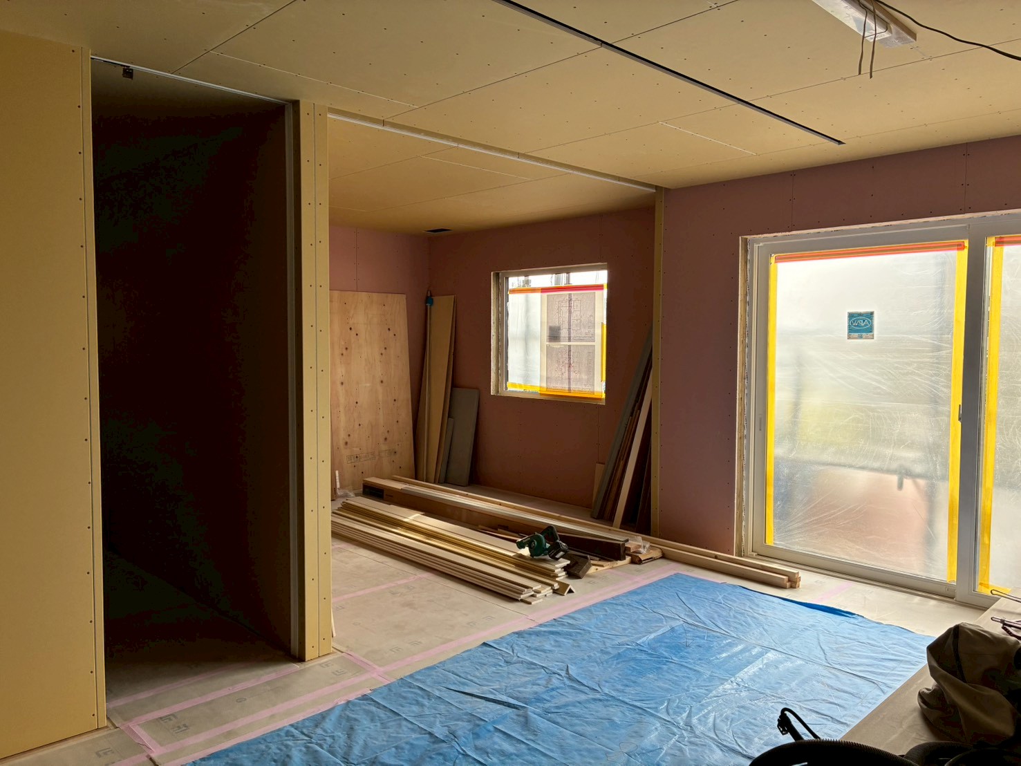 建築工房いろはが施工する注文住宅の建設現場において、内装の石膏ボード施工が完了し、窓やサッシに養生が施され、床にはブルーシートや資材が置かれた家づくりの進捗状況を伝える写真。