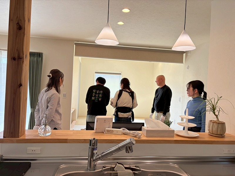 建築工房いろはが開催した完成見学会の様子で、対面キッチン越しに開放的なリビングや小上がりの和室を内覧する来場者と、家づくりのこだわりを共有し合う工務店スタッフのコミュニケーション風景を捉えた写真。
