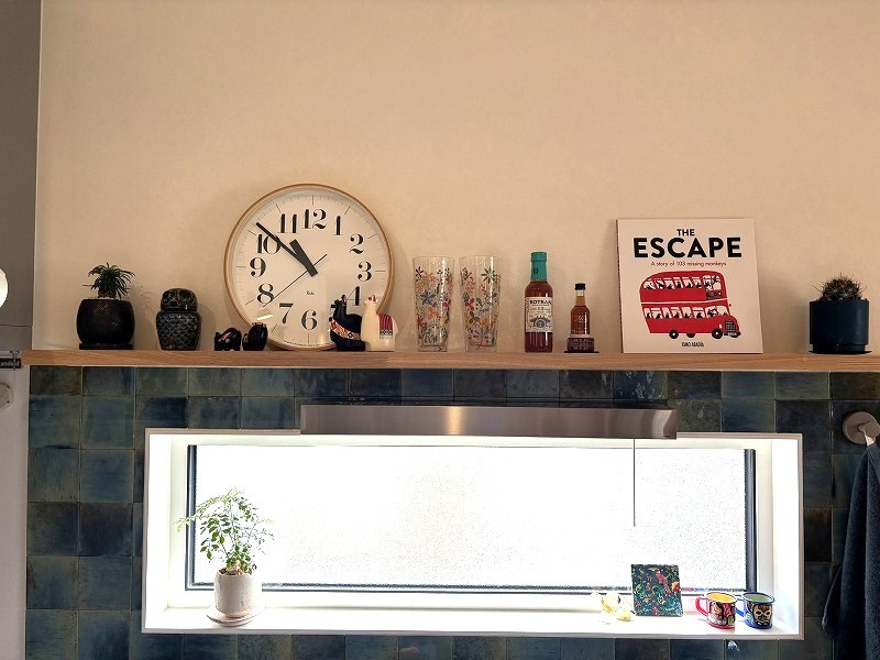 建築工房いろはが手掛けた注文住宅の室内を撮影した写真であり、青いタイルの壁面に設置された飾り棚に、木製の時計や観葉植物、絵本などの雑貨が配置され、明るい窓辺の空間とともにこだわりの家づくりを形にしたインテリアが映し出されています。