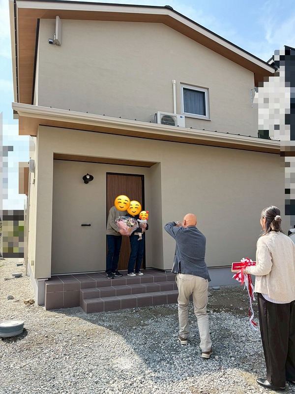 工務店の建築工房いろはが手掛けた注文住宅の引き渡しセレモニーにて、ベージュの外壁が印象的な新築住宅の玄関前で、花束を抱えたご家族をスタッフが記念撮影している、家づくりの感動的な瞬間を収めた写真です。
