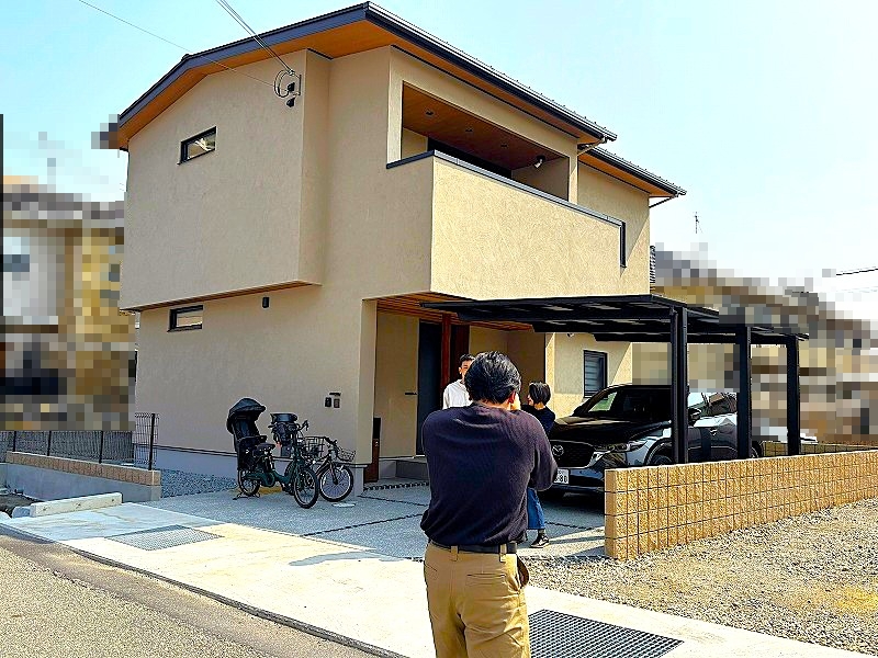 建築工房いろはが手掛けた注文住宅の施工事例であり、落ち着いたベージュの外壁が特徴的な二階建て住宅の外観とともに、黒いカーポートに停まる車両や自転車、家づくりの現場で撮影や確認を行う人々の姿が記録された、地域に根差す工務店によるこだわりの住まいを伝える写真です。
