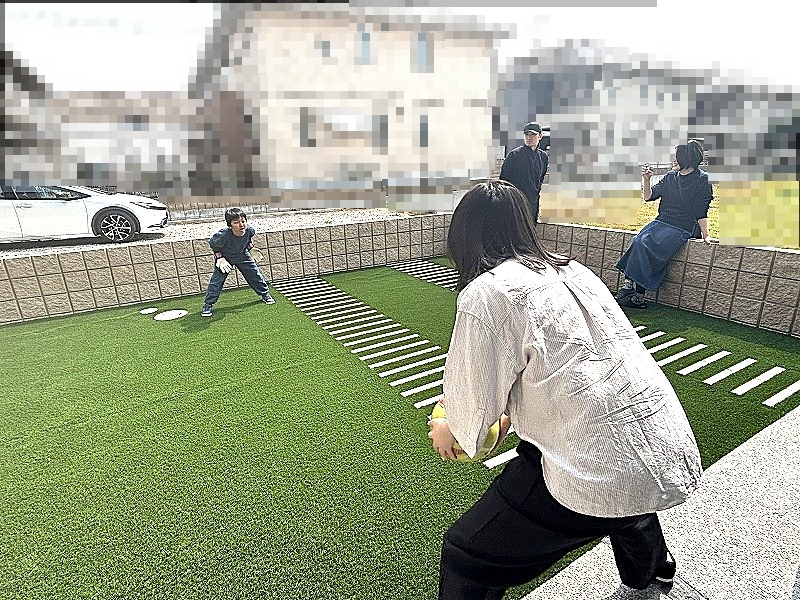 兵庫県の工務店である建築工房いろはが手掛けた注文住宅の庭において、人工芝が敷かれた広々とした空間で家族がボール遊びを楽しむ様子を撮影した写真であり、外構計画を含めた自由な家づくりの実例として、住宅地でのプライベートな庭の活用方法を示しています。