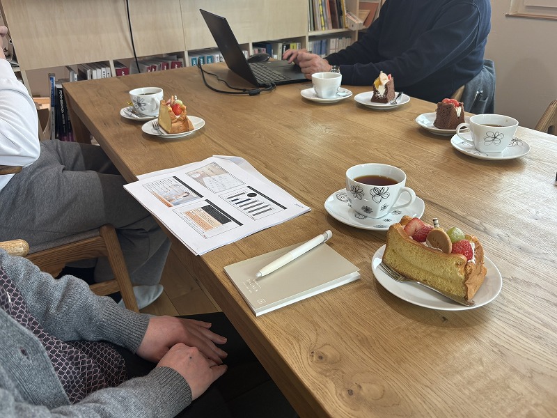 建築工房いろはの家づくり勉強会で、木製テーブルに資料やノートPC、コーヒーとケーキが並び、参加者が注文住宅や資金計画について話し合っている様子を写した写真です。