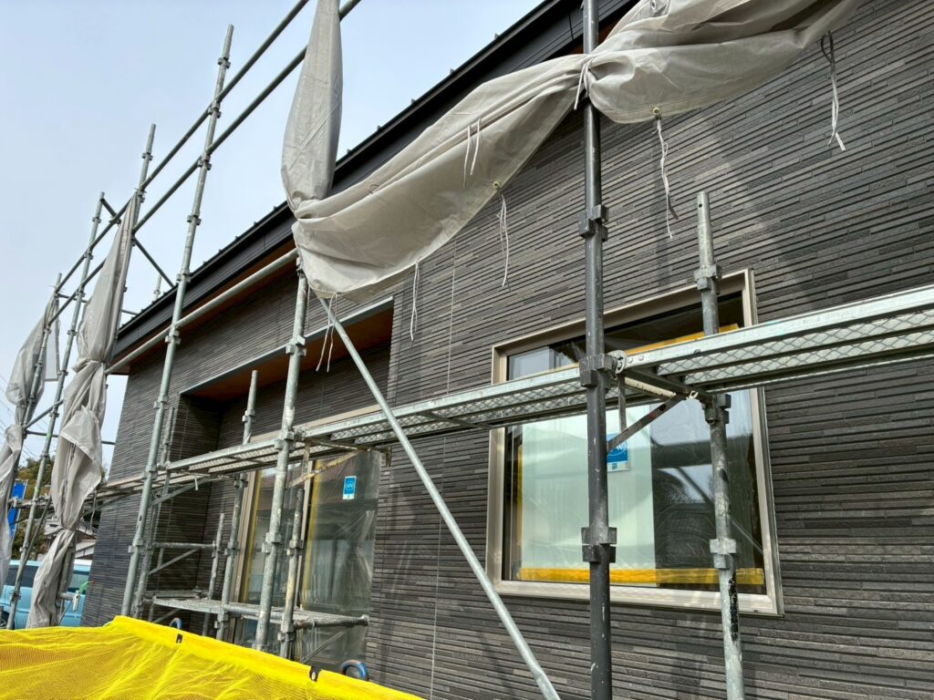 建築工房いろはが施工を手掛ける注文住宅の現場において、黒を基調としたモダンな外壁タイルが施工され、窓周りの養生や足場が組まれた家づくりの進捗状況を伝える写真。