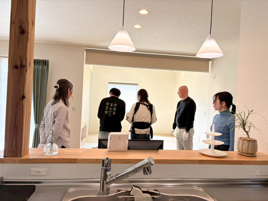 ALTテキスト（社名オプション：建築工房いろは）：
注文住宅の完成見学会において、キッチンカウンター越しに明るいリビングや畳スペースを見学する来場者と、家づくりのこだわりを説明する建築工房いろはのスタッフの様子を捉えた、工務店による内覧会の活気ある雰囲気が伝わる写真です。