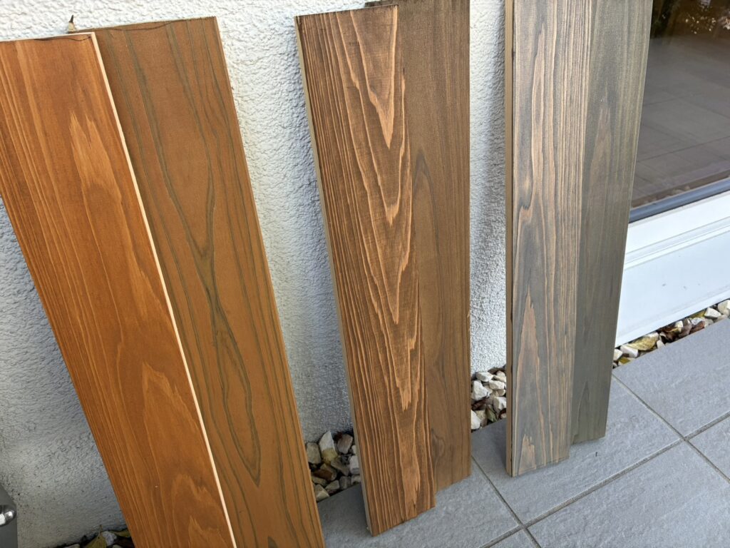 明るい茶色から灰がかった色まで、木目や色調が異なる複数の塗装サンプル材が、白い外壁とグレーのタイル床の間に立てかけられている様子