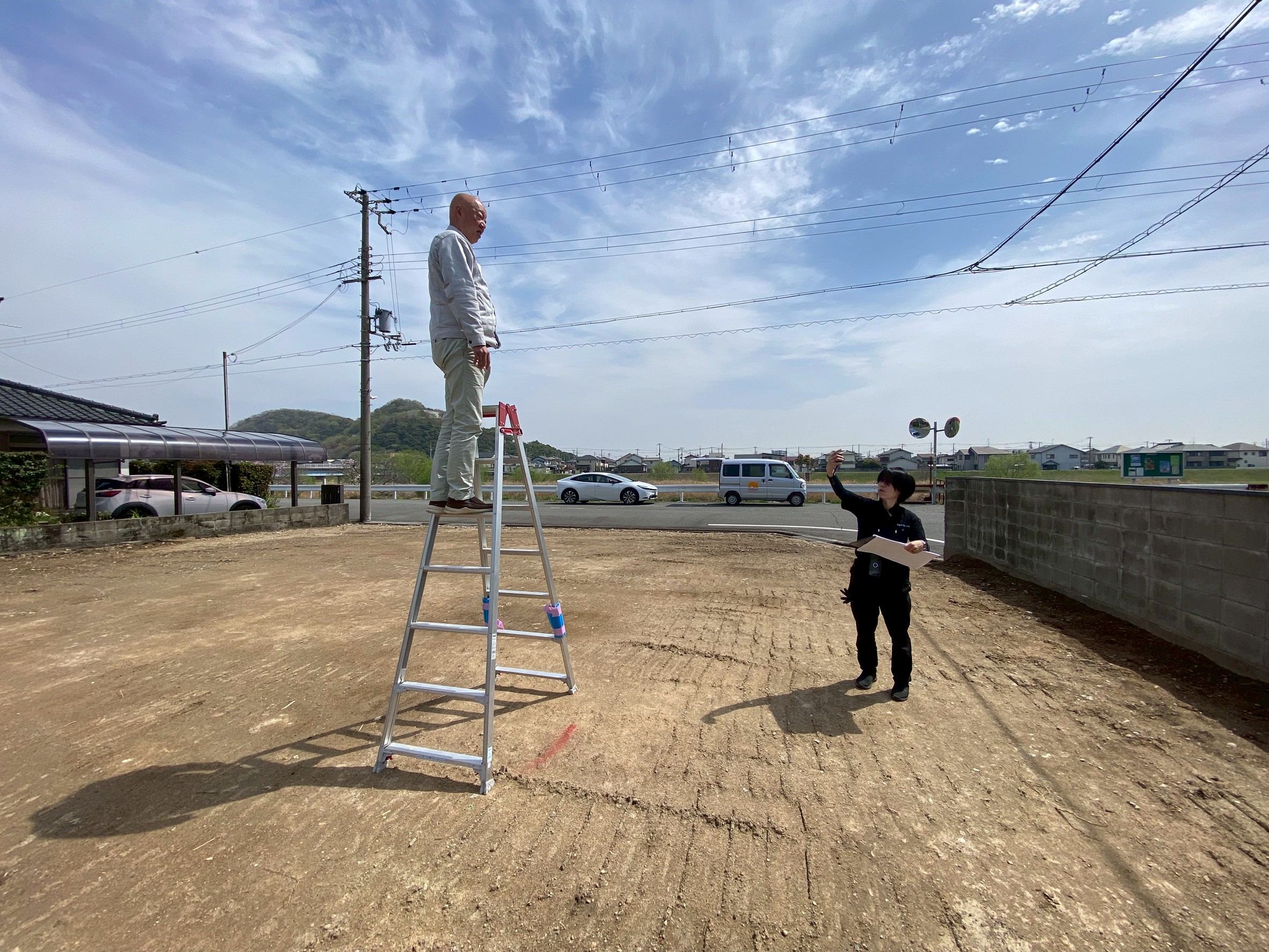 晴天の下、更地となった建築予定地にて、脚立の上に立ち周囲を確認するスタッフと、地上で図面を手に指示や確認を行うスタッフによる、着工前の現地調査の様子。