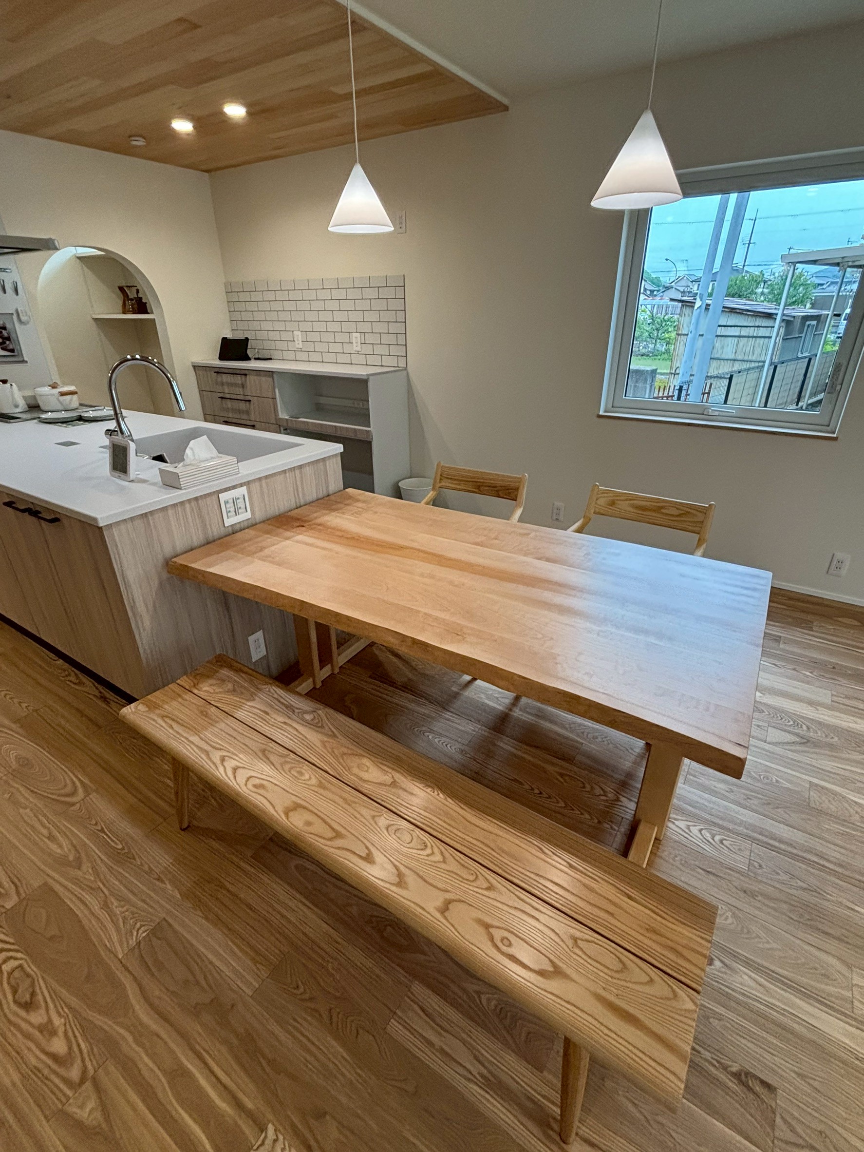 木の温もり溢れる無垢材のダイニングテーブルとベンチが主役のダイニング。折り上げ天井の木目とペンダントライトが調和し、キッチン奥のアール壁のパントリーやタイル仕上げの背面収納がアクセントを添えるナチュラルな住空間。