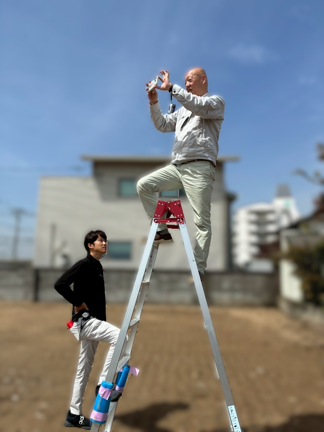 青空の下、更地にて脚立に登り高い位置からスマートフォンで現場撮影を行うスタッフと、下から見守りながら状況を確認するスタッフによる、入念な現地調査の様子。