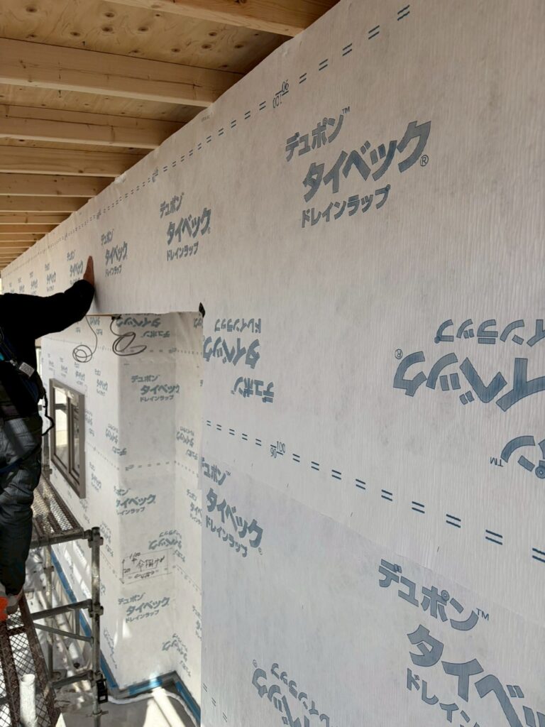 住宅の建築現場にて、職人が外壁下地の透湿防水シート「デュポン タイベック ドレインラップ」を丁寧に施工している様子。シワなく張り込まれたシートが、建物の耐久性と防水性を高める重要な工程を示しています。