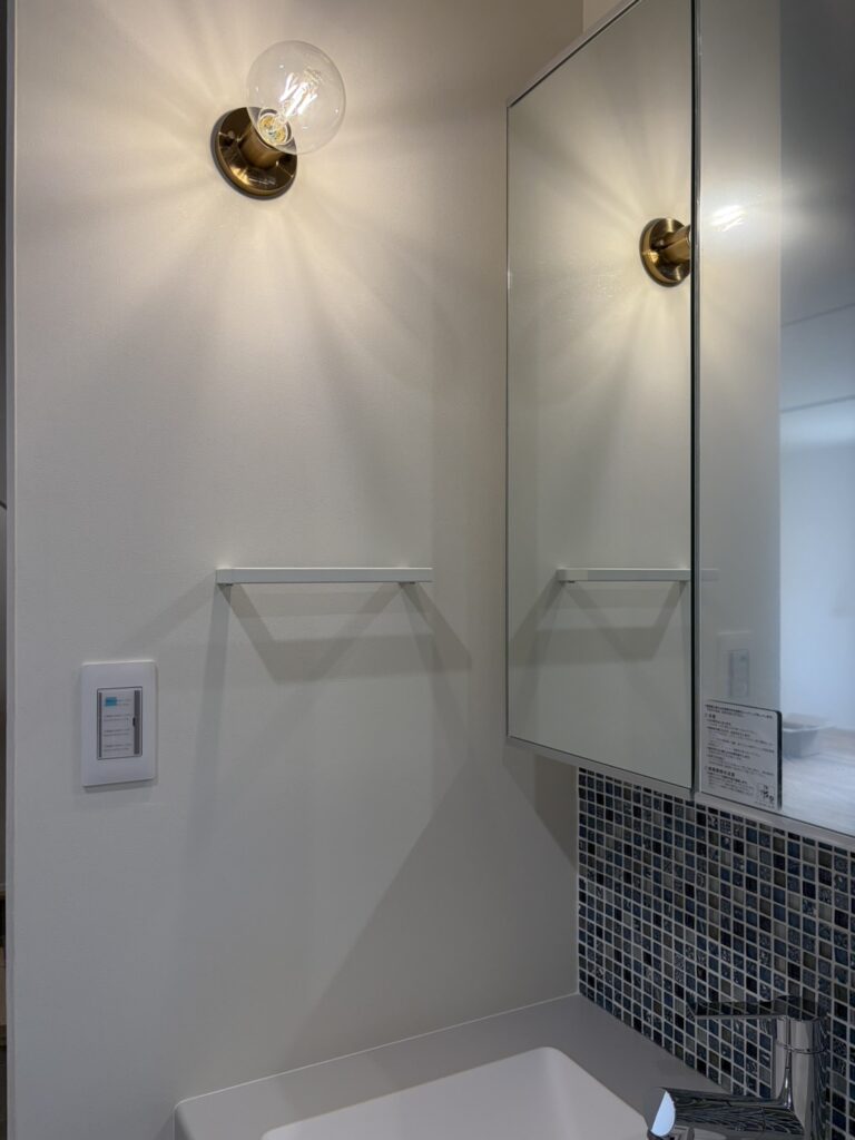 真鍮色のベースがクラシカルな雰囲気を演出するエジソン電球のブラケットライトと、ブルー系のモザイクタイルをあしらった洗面台。シンプルで清潔感のある白い壁に、機能的なミラーキャビネットを組み合わせたおしゃれな洗面空間です。