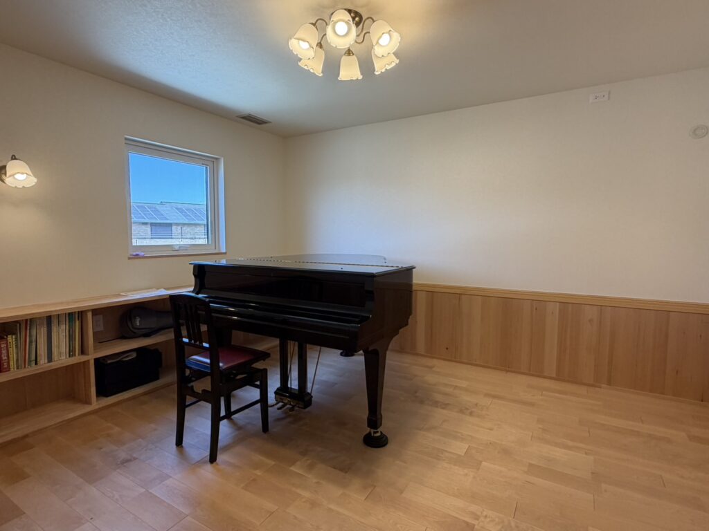無垢材のフローリングと腰壁に囲まれた、木の温もりあふれるピアノ室。中央にグランドピアノが配置され、アンティーク調のシャンデリアとブラケットライトが優しく空間を照らす、防音や音響に配慮した落ち着きのあるレッスンルームです。