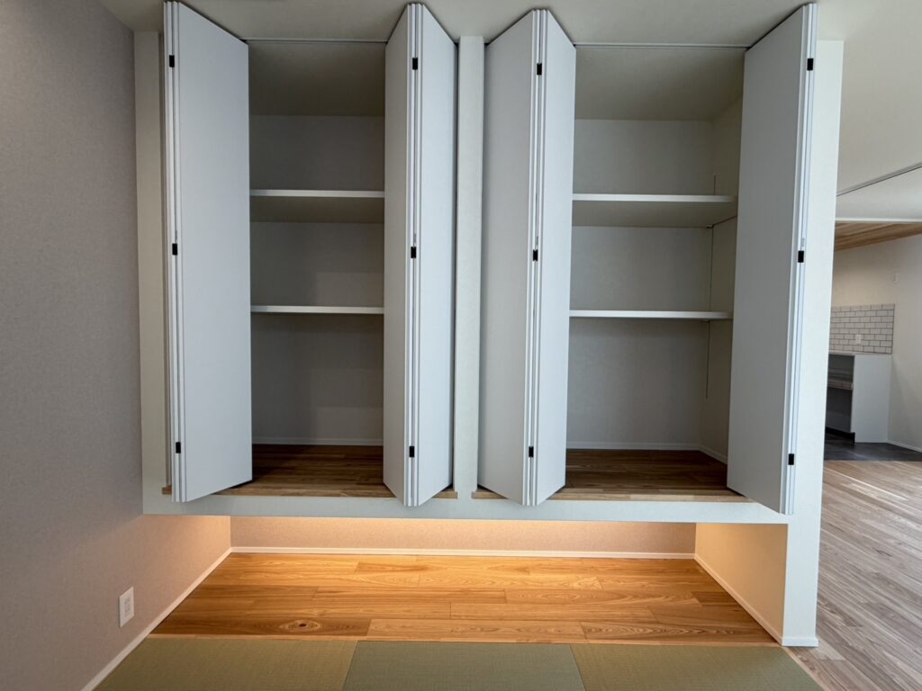 畳コーナーに設けられた、間接照明が足元を優しく照らす吊り押し入れ。白の折れ戸を採用した大容量の収納スペースと、無垢材の質感が調和する、機能的でモダンな和室のデザインです。