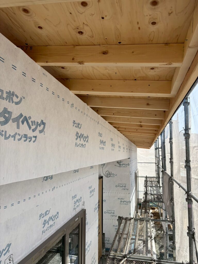 建築現場で、木造住宅の軒天と外壁下地にデュポン タイベック ドレインラップが施工されている様子。足場が組まれ、木材の構造と防水シートの正確な施工を確認できる現場写真です。