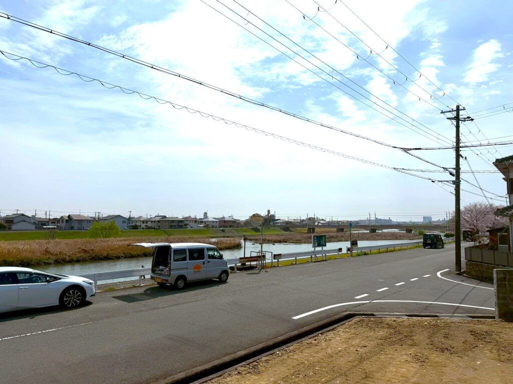 薄曇りの空の下、川沿いに停車するシルバーの作業車と白の乗用車。背景には穏やかな川の流れと対岸の住宅街が広がり、遠くに橋が見える施工現場周辺の風景。