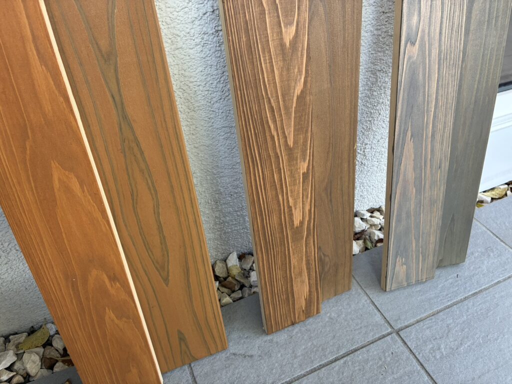 明るい茶色から灰色がかった色まで、木目や色調が異なる複数の塗装サンプル材が、白い外壁とグレーのタイル床の間に立てかけられている様子