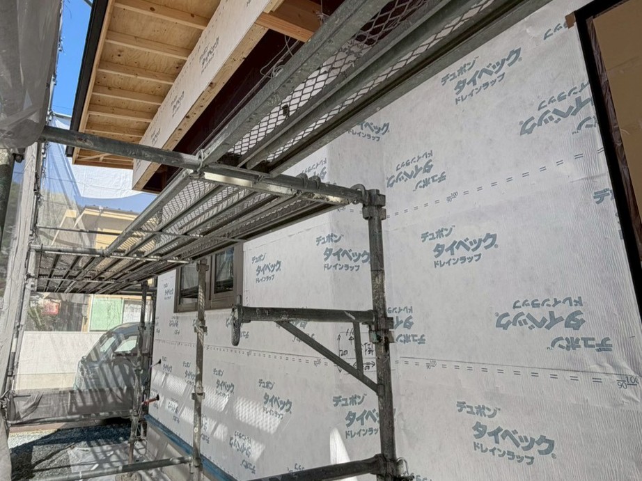 建築現場の足場越しに見える、外壁下地にデュポン タイベック ドレインラップが隙間なく施工された住宅の構造写真。木造住宅の透湿防水シート施工による、高い防水性と通気性の確保を伝えています。