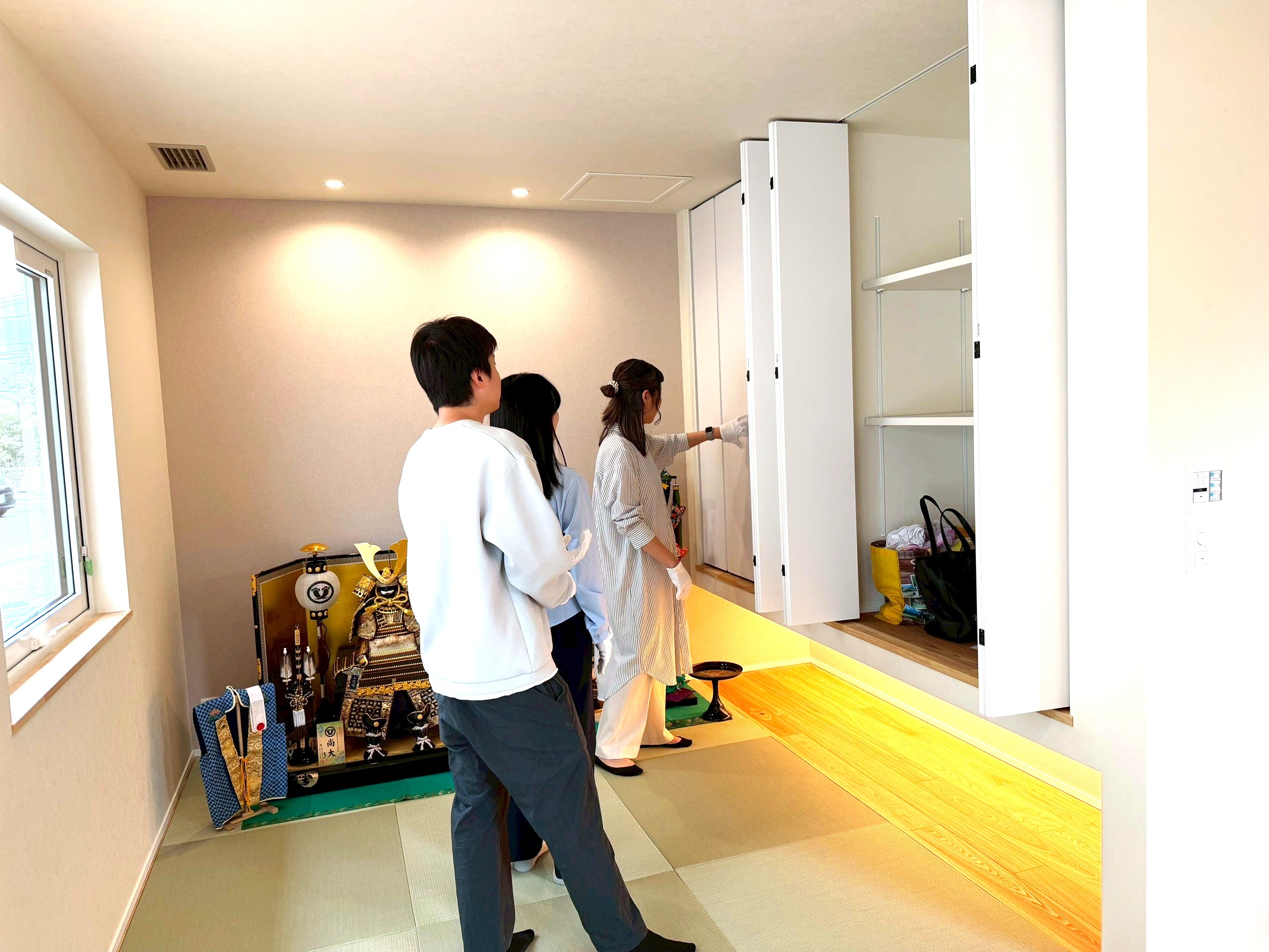 モダンな和室で吊り収納を内覧する施主。畳敷きの空間には五月人形が飾られ、フロアライトの柔らかな間接照明が明るいフローリングを照らす、収納力とデザイン性を両立した和モダンな住まい。
