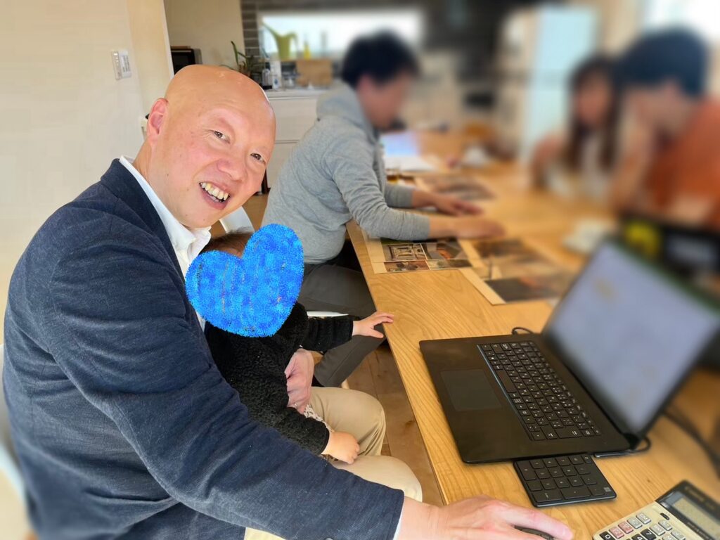 明るい打ち合わせスペースで、ノートパソコンや電卓を使いながら笑顔で家づくりの相談に応じるスタッフ。膝にはお子様を抱いており、お子様連れでも安心して相談できる様子が伺えます。