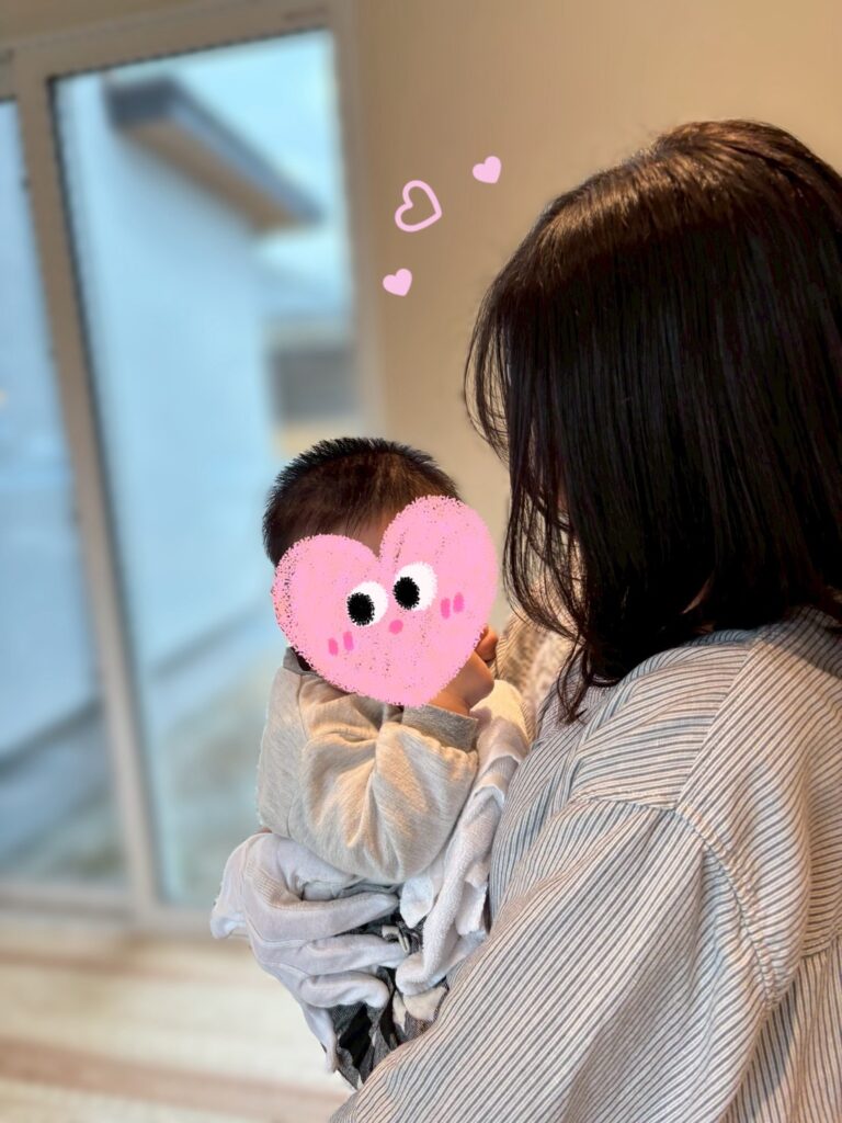 明るい光が差し込む大きなサッシを背景に、赤ちゃんを愛おしそうに抱っこするお母様。温かみのある室内空間で、ご家族が笑顔で心地よく過ごせる家づくりの様子が伝わります。