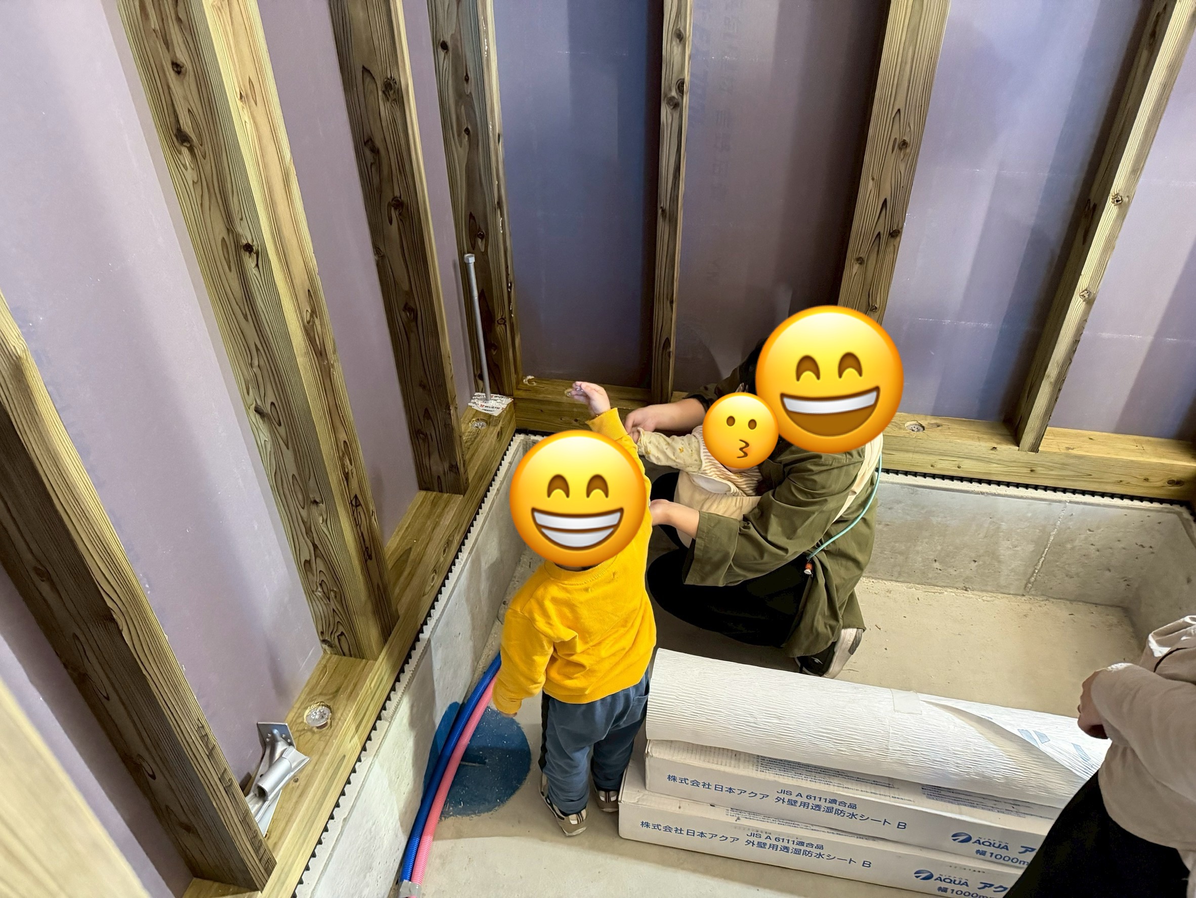 建築中の木造住宅の内部で、母親に抱かれた赤ちゃんと、黄色の服を着た子供が、構造材や配管を興味深そうに見つめる様子。床には防水シートや配管材が置かれている。
