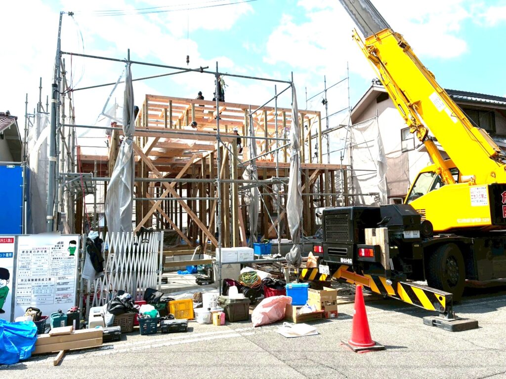 青空の下で進む木造住宅の上棟作業。組み上がった構造体と、現場で稼働する大型の黄色い移動式クレーン車、整理整頓された資材や看板が、活気ある建築現場の様子を伝えています。