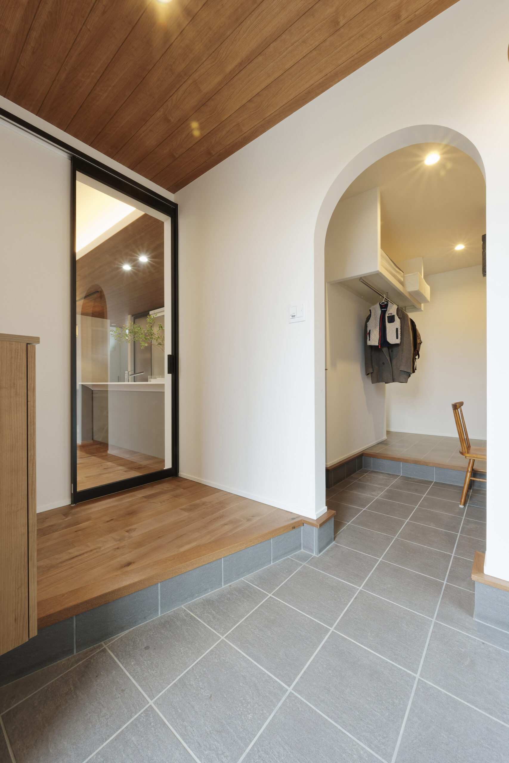 木目調の天井が温かみを添える、開放的なタイル床の玄関ホール。アーチ状の垂れ壁がアクセントのファミリークローゼットへ直接つながる動線や、リビングの様子が見えるガラス扉を採用した、明るくモダンな空間設計です。