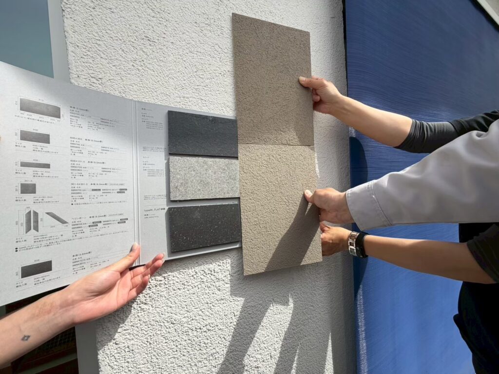 木製デッキのテラスにて、建築担当者と施主が実際の外壁サンプルを壁に当て、質感や色合いを確認しながら打ち合わせを行う様子。