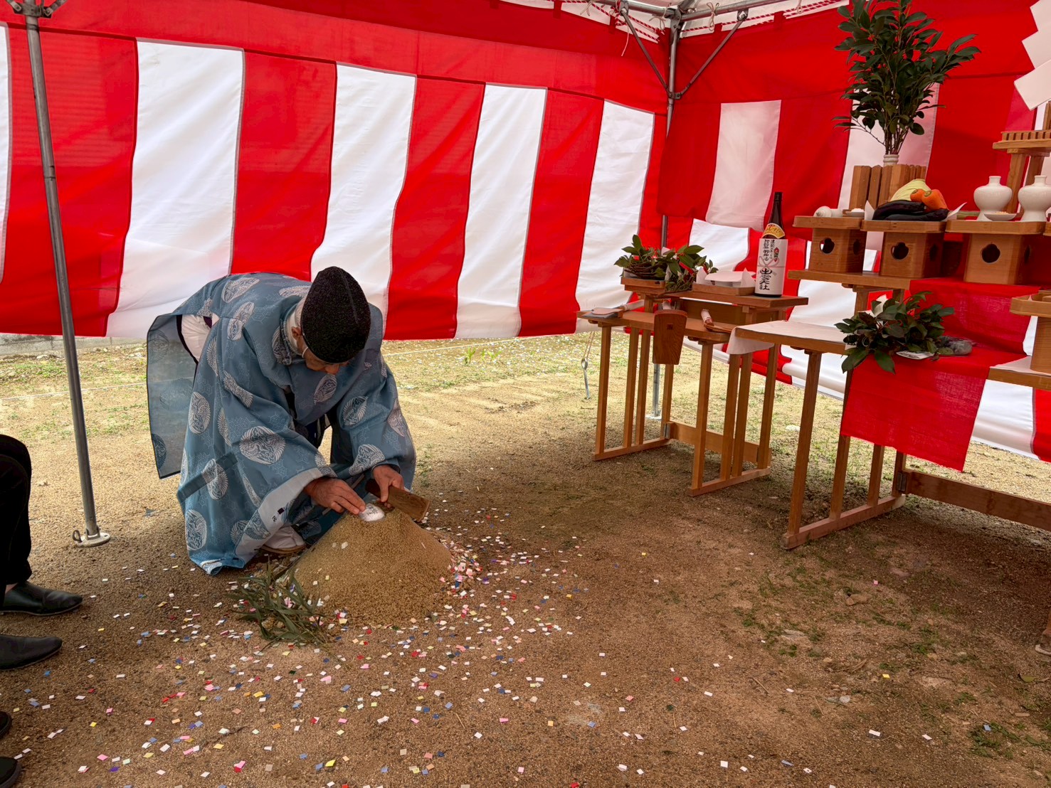 紅白幕が張られた地鎮祭の会場で、神職が砂盛に対して穿初（うがちぞめ）の儀を行う様子。傍らにはお供え物が並ぶ祭壇が設置され、厳かな家づくりの儀式が執り行われています。