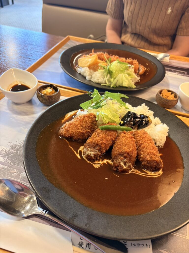 濃い色のカレーソースに、揚げたてのカキフライが4つと素揚げした茄子、彩りの良い野菜が添えられたプレート料理。落ち着いた色合いの皿に盛り付けられ、付け合わせの小鉢も並ぶ食卓の様子。