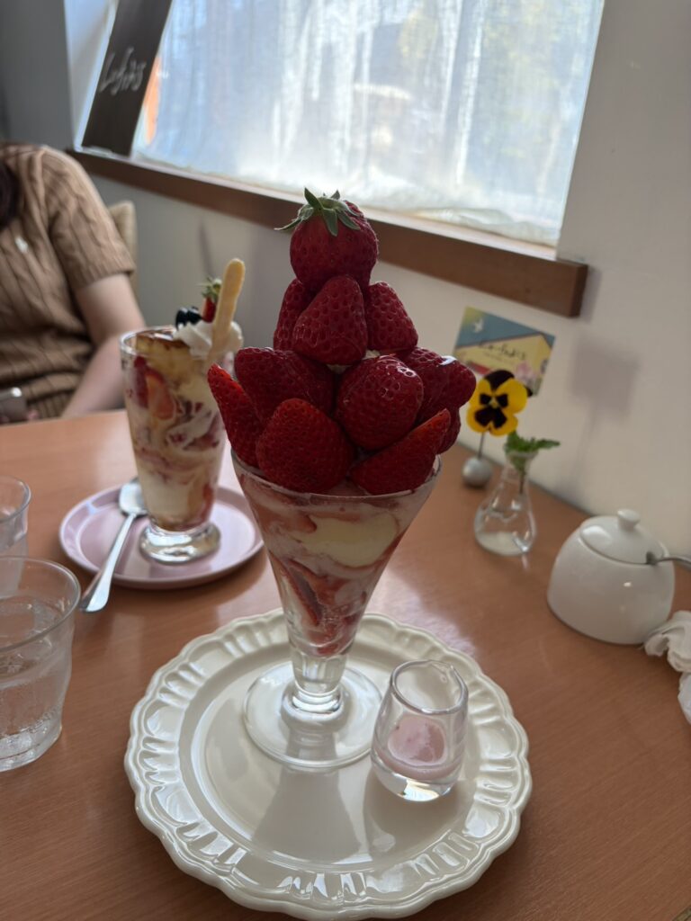 テーブルに並んだ二つの華やかなイチゴのパフェ。手前にはイチゴが山盛りに積み上げられたパフェが、奥には別のデザートが置かれ、明るい窓辺のカフェで過ごすゆったりとしたティータイムの様子。
