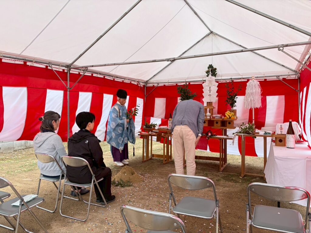 紅白幕が張られたテント内で、神職に見守られながら施主が祭壇に向かって一礼する地鎮祭の様子。祭壇には玉串や果物などのお供え物が整えられ、厳かな雰囲気の中で工事の安全を祈願しています。
