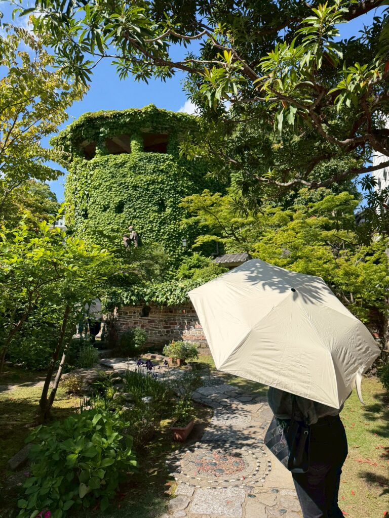 蔦（つた）などの緑に覆われた円筒形の建物と、手入れされた庭園へと続く石畳の小道。