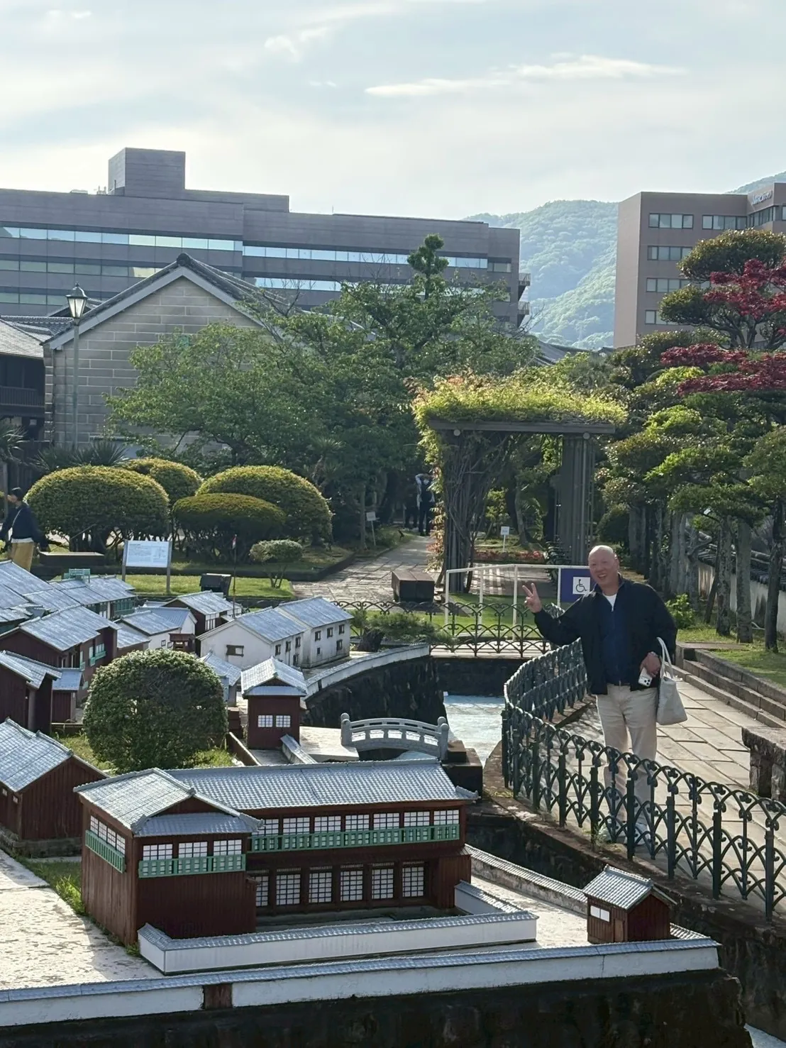 ミニチュアで再現された出島オランダ商館跡の歴史的な和洋折衷建築の街並みと、その横に立つ笑顔でピースサインをする男性