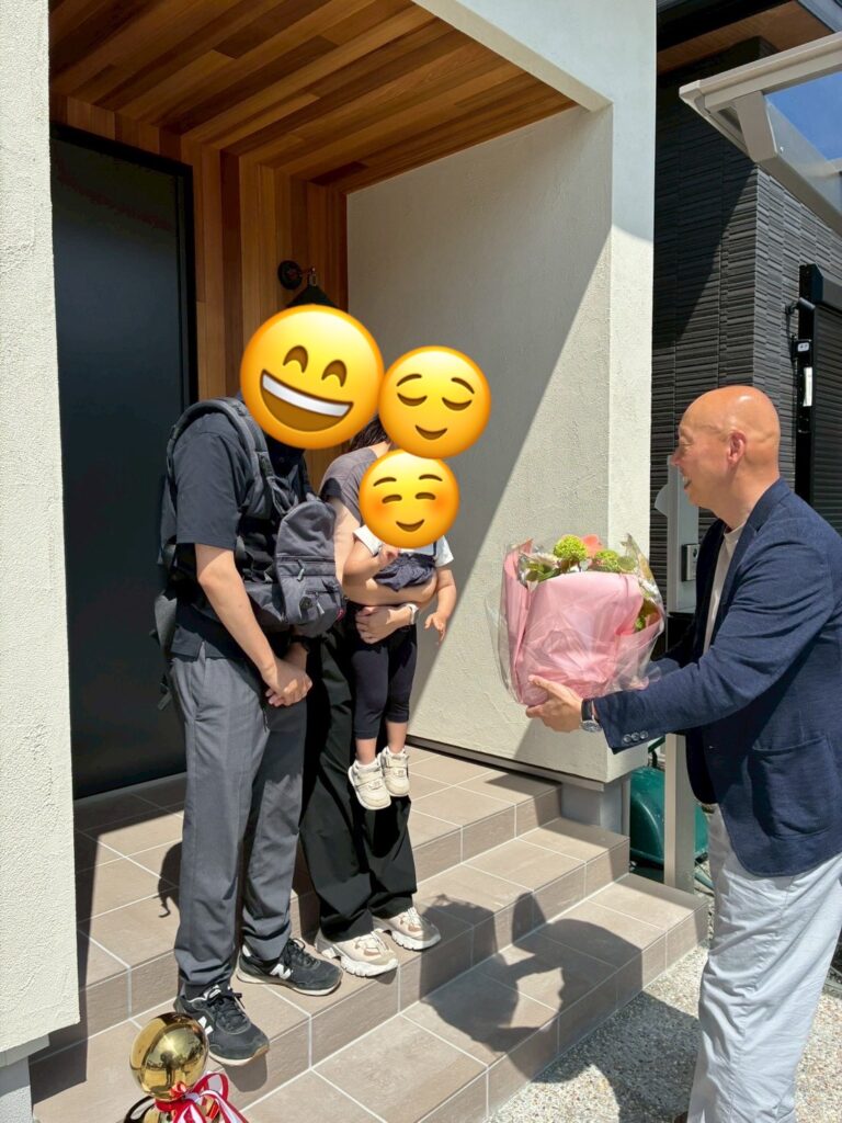 木目調のアクセントパネルが温かみを添える玄関先で、完成を祝して花束が贈呈される、ご家族と工務店担当者の記念すべき引き渡し風景。
