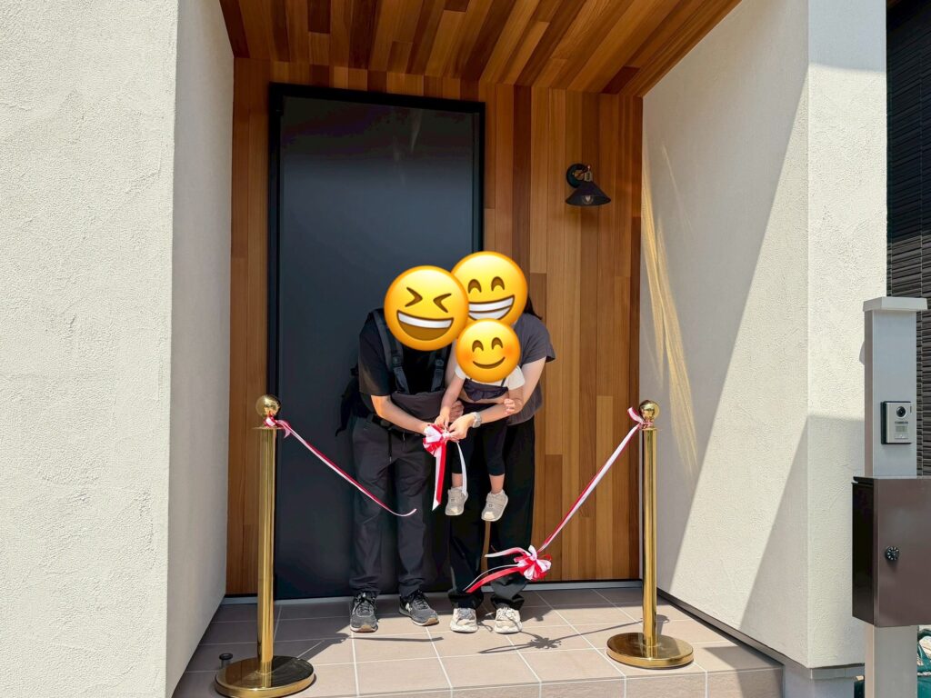 木目調のアクセントウォールと黒い玄関ドアがモダンな印象を与える玄関ポーチにて、ご家族がテープカットを行い完成を祝う様子。